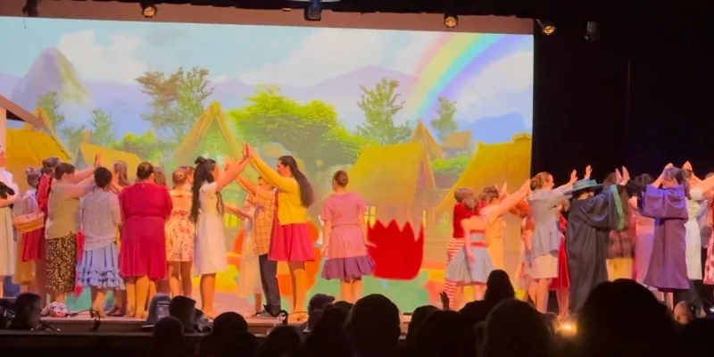A scene from a live theater performance. A group of actors are standing on stage in front of a projected backdrop of a village with rainbows in the sky.